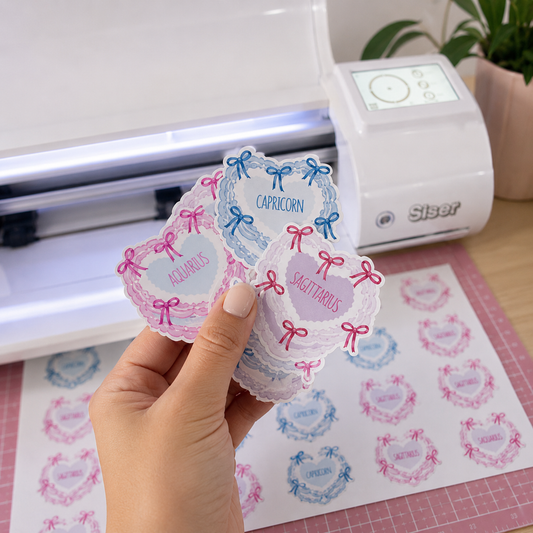 A hand holds a stack of decorative die-cut stickers featuring pastel pink and blue heart-shaped frames with bows and text labels like 'ACEURALS', 'CAPICORN', and 'SHIT TABBARS'. A white Siser cutting machine sits in the background on a wooden desk, with a sheet of matching stickers on a pink cutting mat in front.