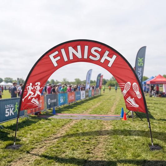 Large red custom printed finish line event arch at an outdoor 5K running race with barriers, flags and event tents.